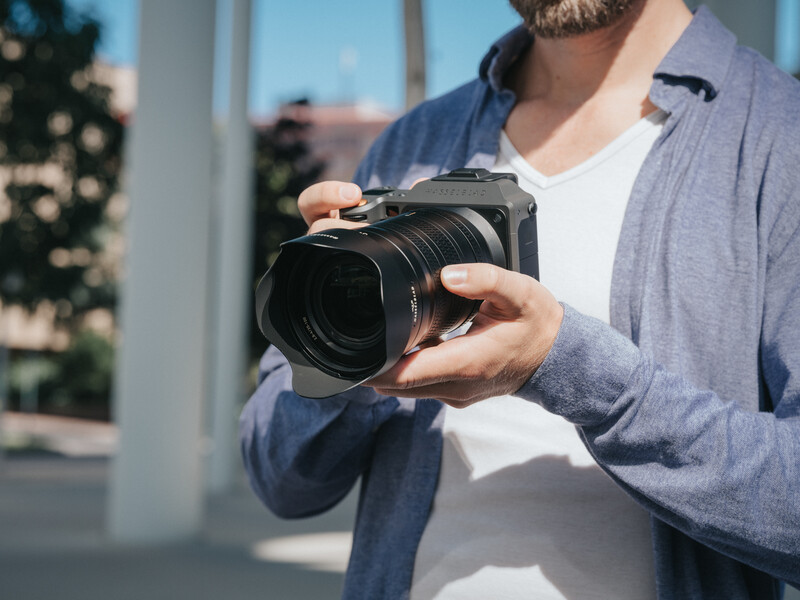 Photographer holding a medium format camera with a large lens, positioned near a bright window with natural outdoor light.
