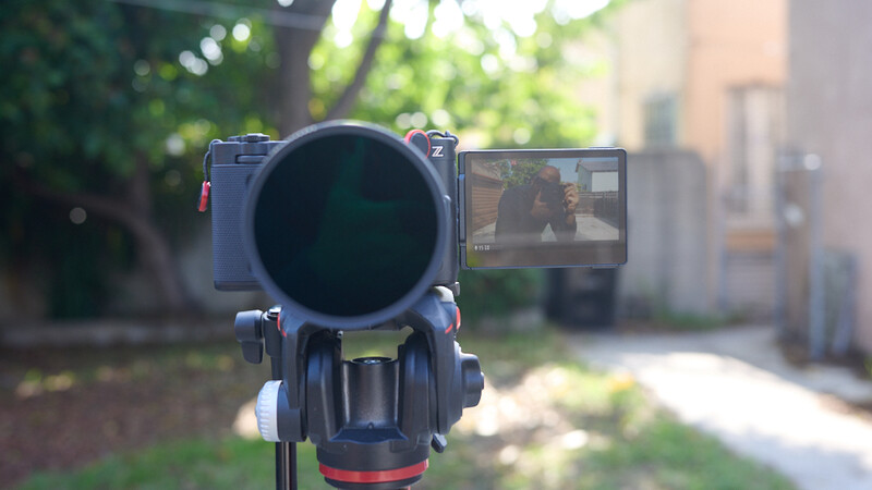 Video camera mounted on tripod with LCD monitor displaying live footage in an urban garden setting.