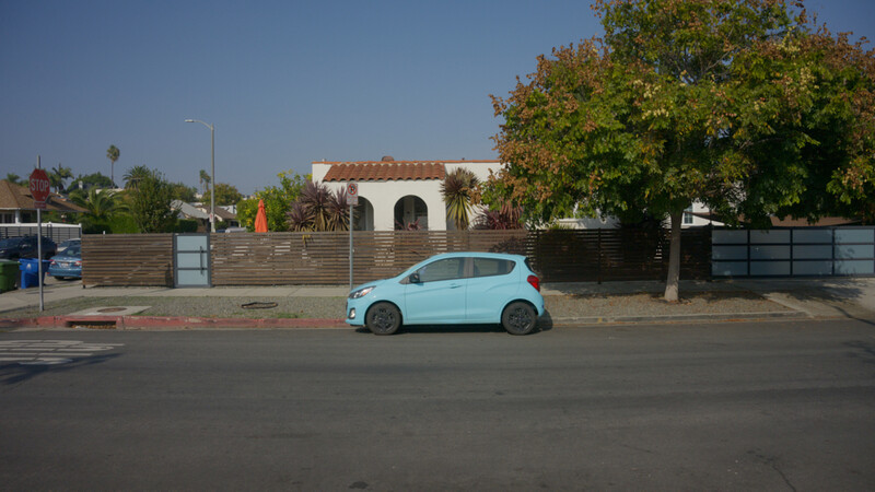 Light blue compact car parked on street beside residential property with white building and wooden fence.