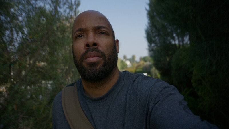 Man with beard looking directly at camera outdoors with evergreen trees in background.