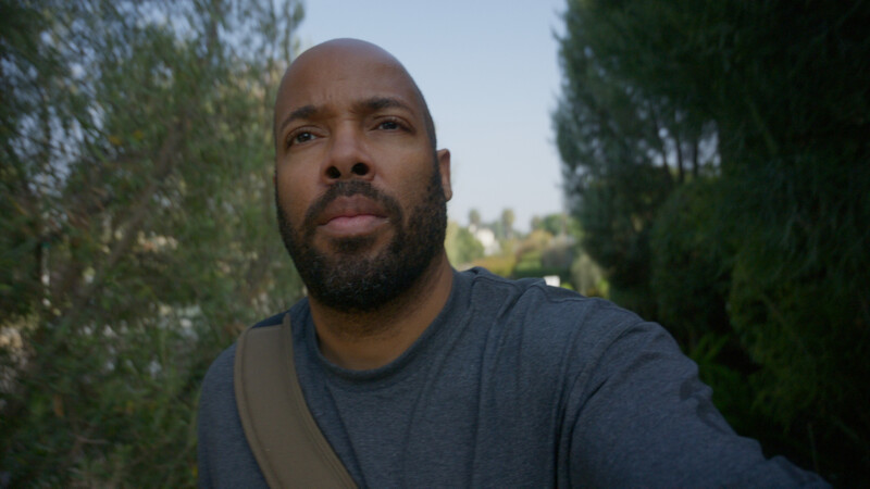 Man with beard wearing dark blue t-shirt, photographed outdoors with evergreen trees and bright daylight in background.