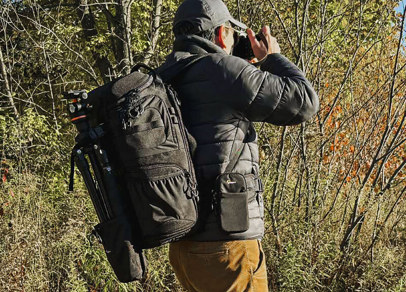 Photographer wearing a dark backpack and mustard-colored pants, holding a camera to their face while standing in a wooded area with sparse vegetation.
