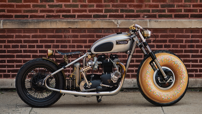 Custom bobber motorcycle with cream tank and gold front wheel against brick wall.
