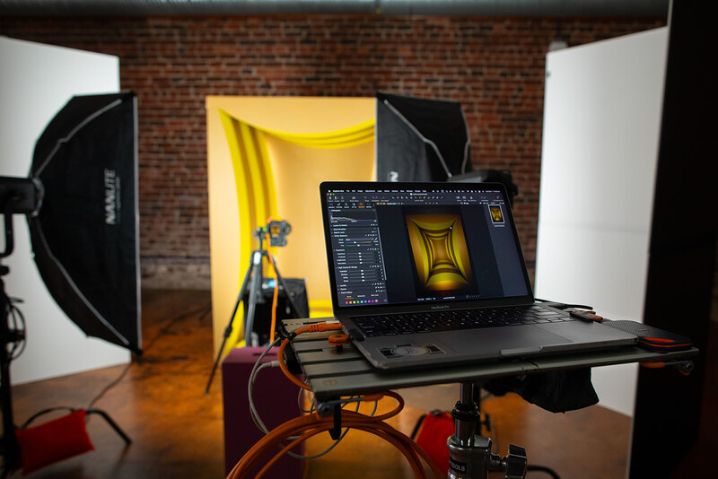 Professional photography studio setup with laptop displaying editing software, professional lighting equipment, and yellow backdrop.