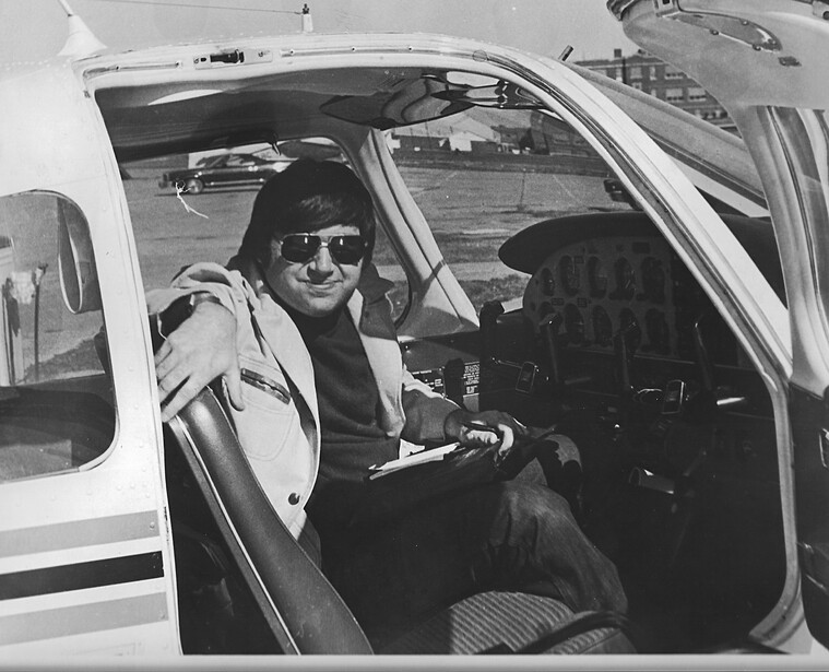 Man wearing sunglasses and light jacket sits in driver's seat of white car, looking toward camera.
