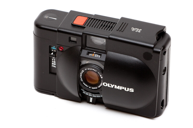 Olympus XA compact rangefinder camera with black body and orange flash button against white background.