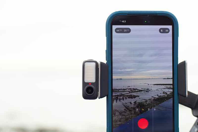 Smartphone mounted on tripod displaying coastal landscape in camera app with red record button visible.