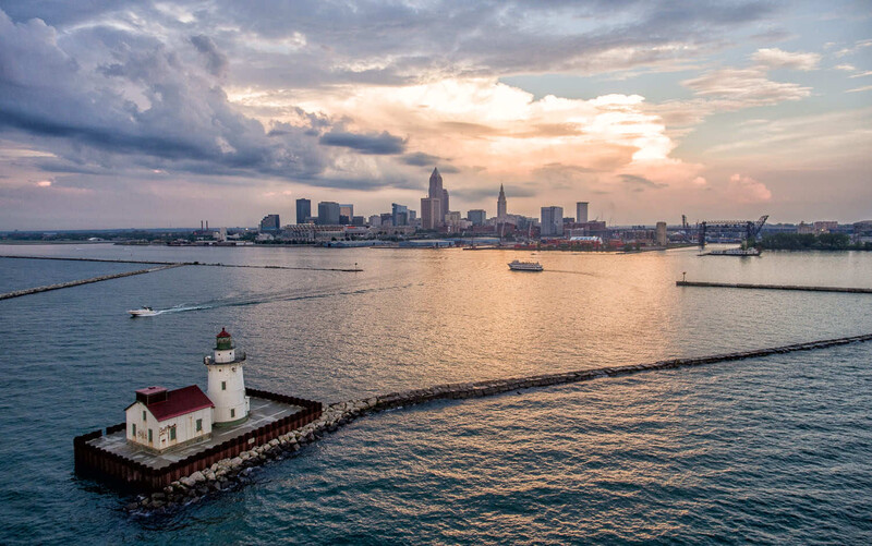 Red-roofed lighthouse on a breakwater with a city skyline and dramatic clouds at sunset across the water.