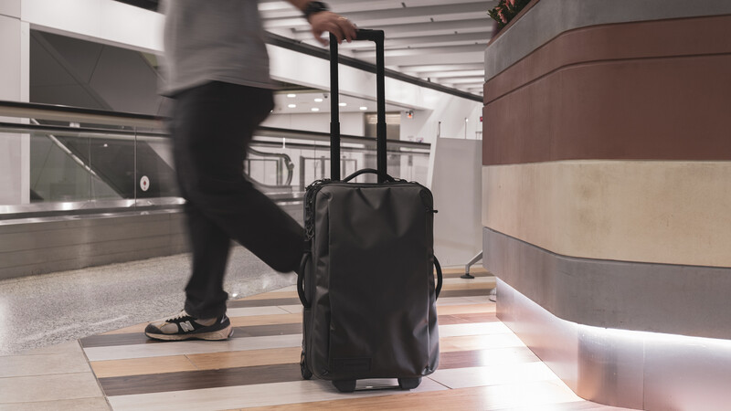 Blurred figure pulling a black rolling suitcase through a modern airport terminal hallway.