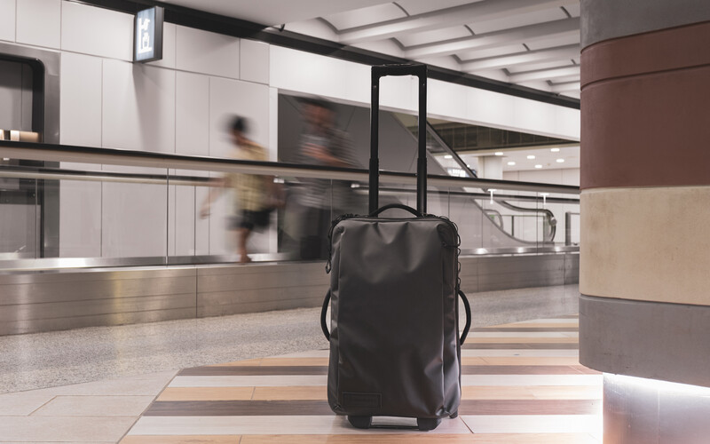 Black rolling suitcase standing alone in a modern airport terminal with blurred travelers passing in the background.
