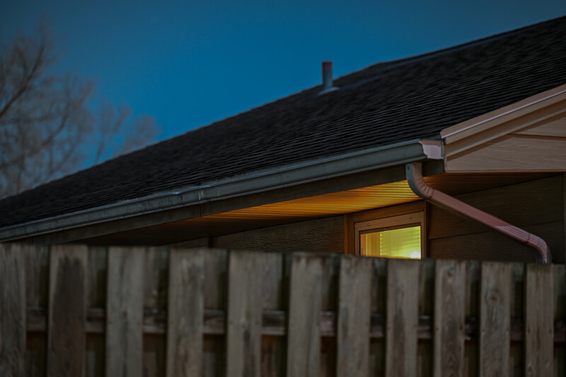 Wooden fence in foreground with illuminated house window and dark roofline against twilight sky.