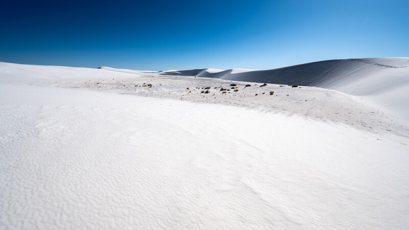 White sand dunes with ripple patterns under clear blue sky, dark volcanic rocks scattered across the ridge.