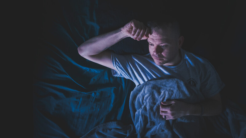 Man in bed at night with hand to head, lit by cool blue light from bedside lamp.