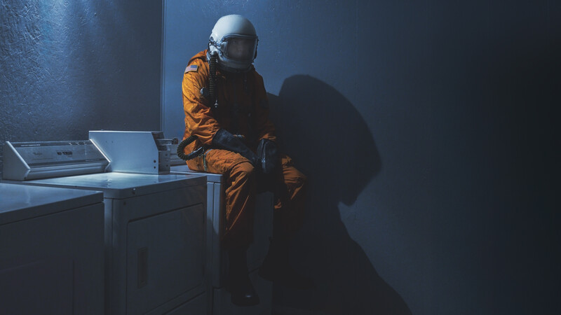 Astronaut in orange suit sitting at desk with laptop in dimly lit room.