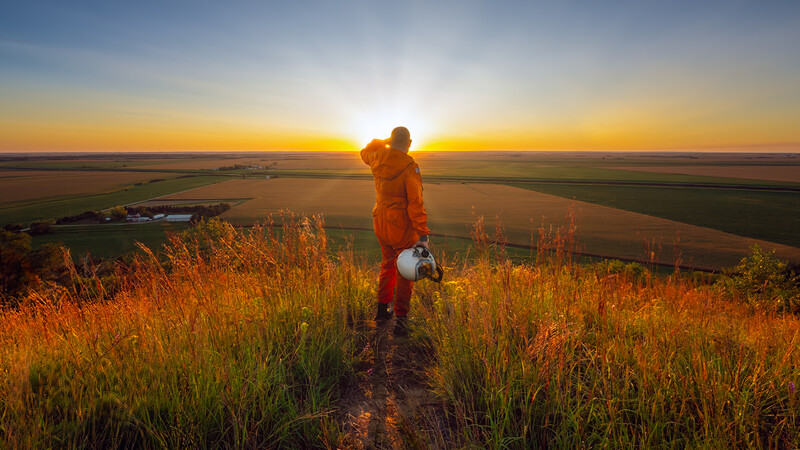 Hiker in orange jacket stands on hilltop overlooking vast farmland at golden hour sunset.