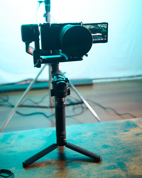 Professional cinema camera mounted on a compact tripod with articulated arm, positioned on a turquoise textured surface against a light blue background.