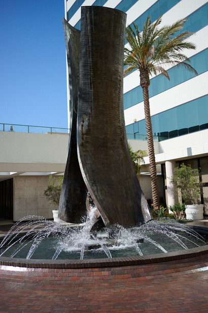 Massive tree trunk with flared base in modern courtyard plaza with striped building facade and palm tree.