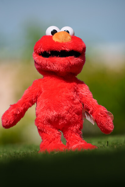 Red Elmo puppet standing outdoors with blurred green and blue background.