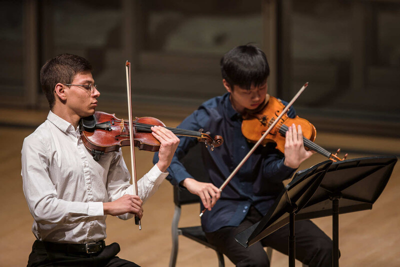 Two musicians seated side-by-side playing violins during a performance.