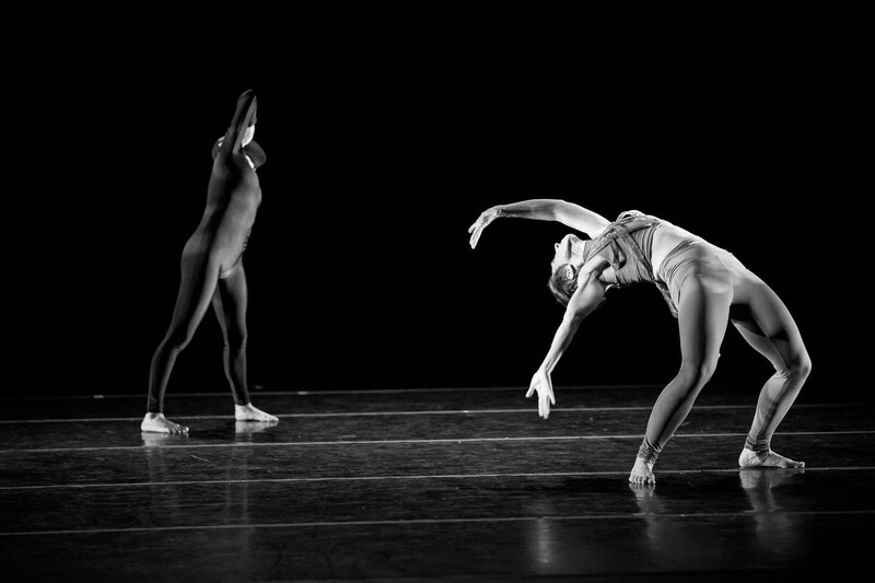Two dancers in contrasting poses on a dark stage floor against a black background.