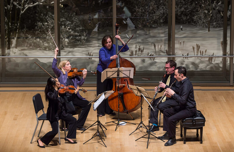 Four classical musicians performing in a modern concert hall with large windows overlooking a landscape.