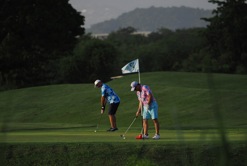 Two young golfers on a putting green with flag stick, mountains visible in misty background.