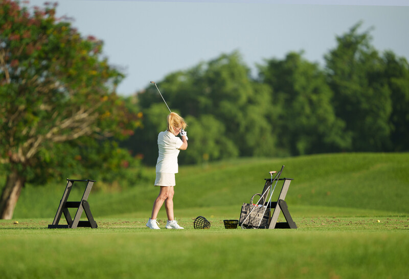 Female golfer mid-swing on a practice range with organized equipment stations and lush green fairway.