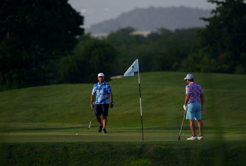 Two golfers on a green with a flagstick, mountains visible in the distant background.