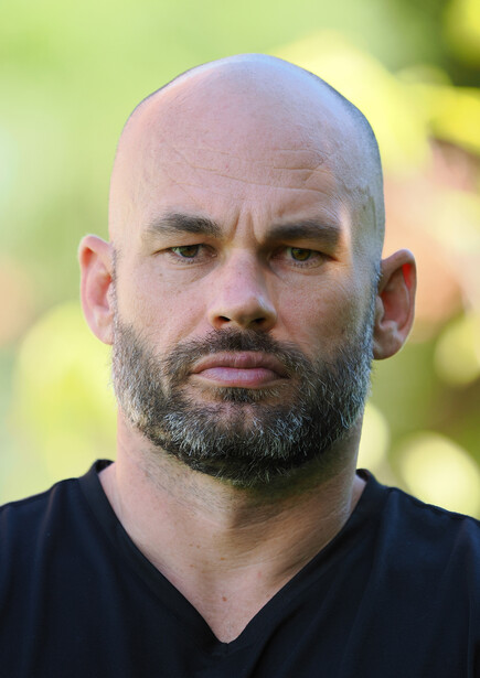 Bald man with full beard wearing black v-neck shirt against blurred green background.