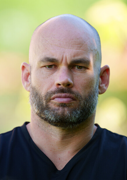 Bald man with beard wearing black v-neck shirt against blurred green and yellow background.
