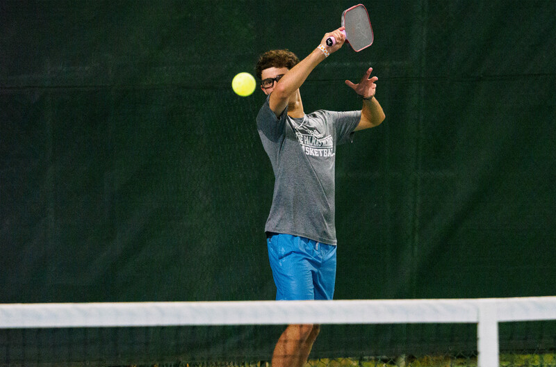 Male tennis player serving overhead on outdoor court with green backdrop.