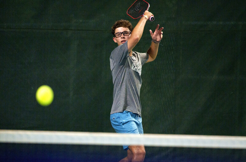 Player in glasses and casual attire making an overhead catch on an outdoor tennis court.
