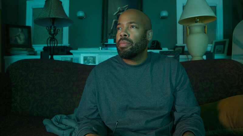 Man sitting on dark couch looking upward in teal-lit interior space.