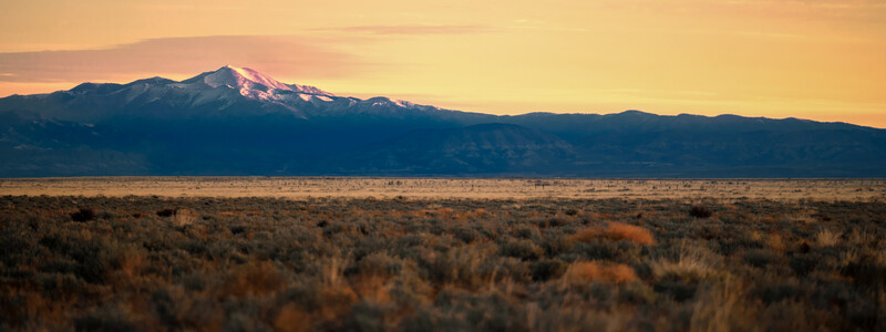 Snow-capped volcanic mountain at sunrise above high desert scrubland.