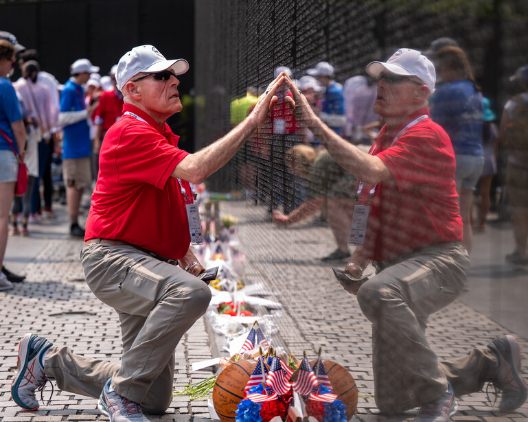 Two men in red shirts kneel facing each other with hands raised in celebration beside memorial flowers at a brick wall.