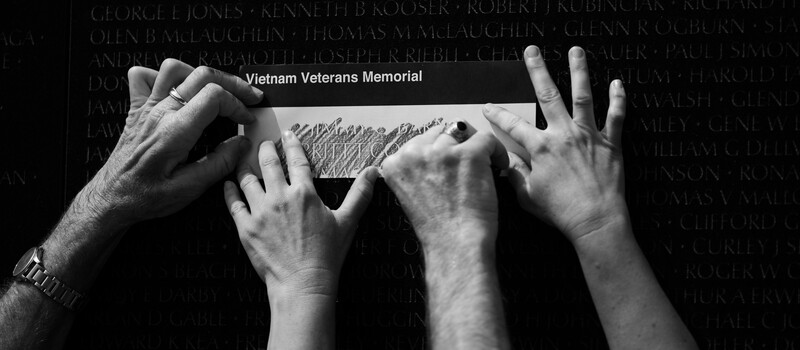 Multiple hands making rubbings on the Vietnam Veterans Memorial wall with names engraved behind.