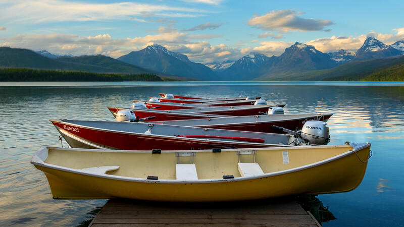 Yellow, red, and white boats moored in calm alpine lake water with snow-capped mountains and forested shoreline in background.
