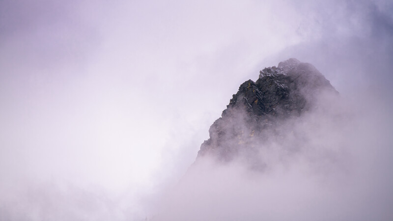 Rocky mountain peak emerging through dense fog and mist in soft purple-gray tones.
