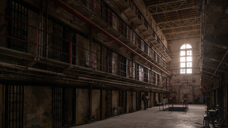 Long corridor of prison cells with barred doors lining both sides, lit by a distant window.