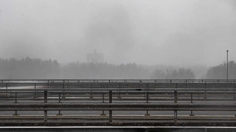 Multi-lane highway bridge stretching into heavy fog with evergreen forest silhouettes in misty background.