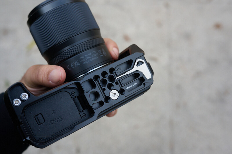 Photographer's hand holding a mirrorless camera body with attached lens and L-bracket, shown from above against a concrete background.
