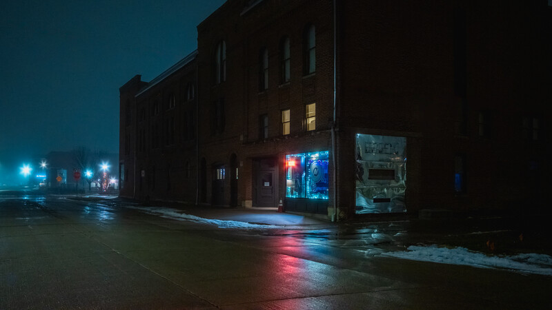 Dark brick building at night with illuminated storefront windows and wet pavement reflecting neon lights.