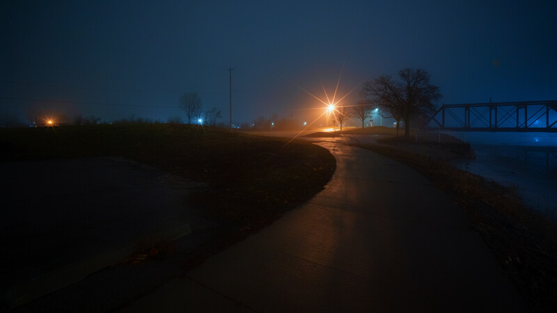 Wet rural road at dusk with street lights and a bridge in the distance under darkening sky.