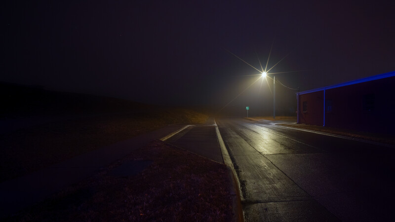Empty industrial parking area illuminated by a single street lamp at night, with dark sky and a blue-roofed structure visible on the right.