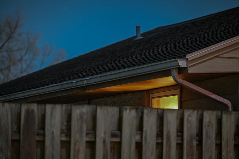 Residential house exterior at dusk with warm interior lighting glowing through a window behind a weathered wooden fence.