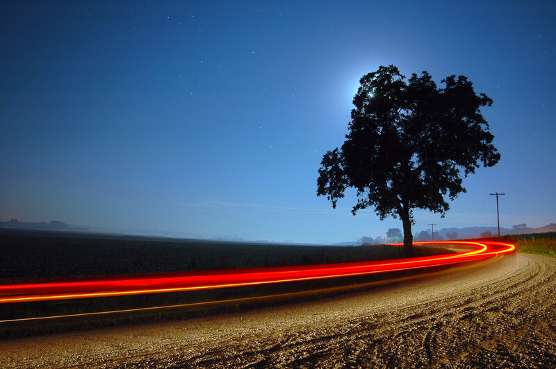 Long-exposure photograph of red vehicle light trails streaking across a rural road at dusk, with a solitary tree silhouetted against a blue sky.