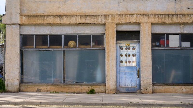 Weathered storefront with boarded windows and blue door, concrete facade stained with age and moss growth.