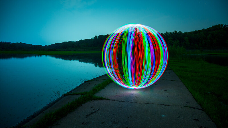 Glowing multicolored sphere with vertical light streaks sits on a concrete dock extending into a still lake at dusk.
