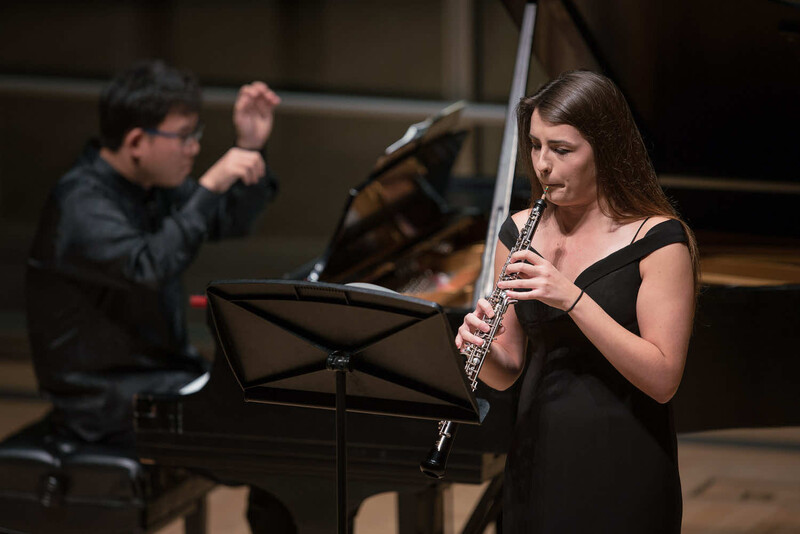 Female flute player performing in concert while conductor directs in background.