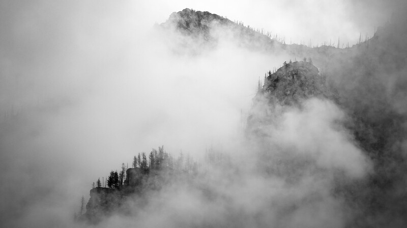 Forested mountain peaks partially obscured by layered fog and mist in a monochromatic landscape.